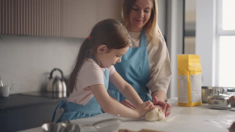 Neugieriges-Kleines-Mädchen-Hilft-Mutter-Beim-Kochen-In-Der-Küche-Und-Lernt,-Teig-Auf-Dem-Tisch-Zu-Kneten-Und-Kuchen-Zuzubereiten